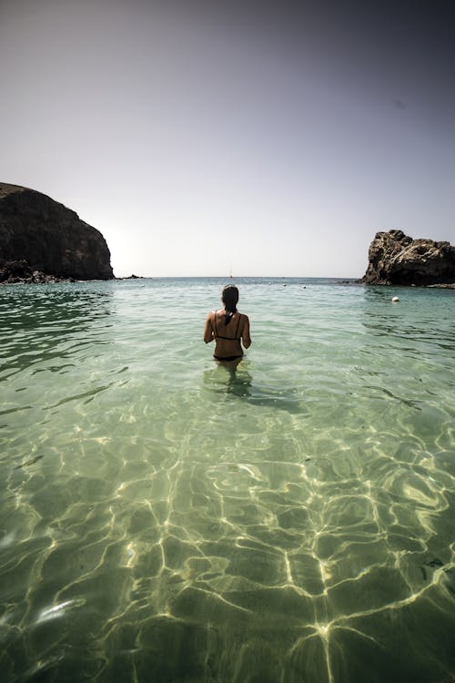 Back view unrecognizable female in swimsuit chilling in crystal clear seawater near rocks and enjoying sunny weather