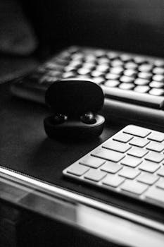 Black and white photo of wireless earphones on a desk with a keyboard.