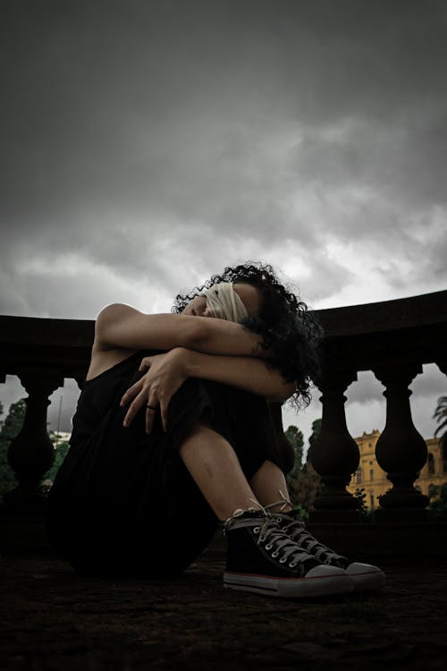 Full body of anonymous depressed lady with curly hair and sitting on balcony hugging knees with covered eyes on overcast day