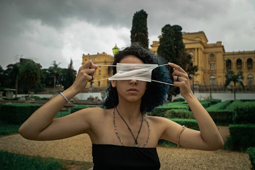 Serious young ethnic female with curly hair in light top covering eyes with medical mask while standing in city park against cloudy sky