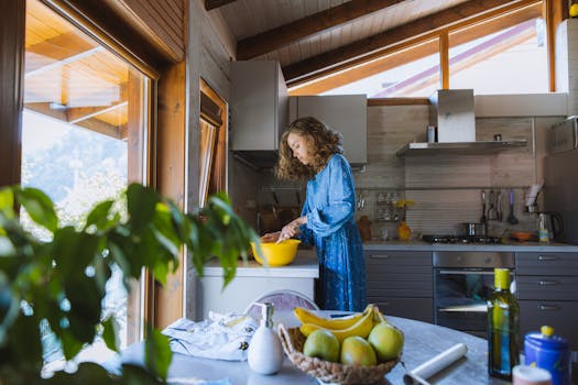 A woman with curly hair prepares a meal in a sunlit kitchen, surrounded by fresh fruits.