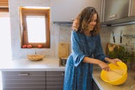 穿着蓝色连衣裙的微笑女人在舒适的厨房里把面团和透过窗户的阳光混合在一起