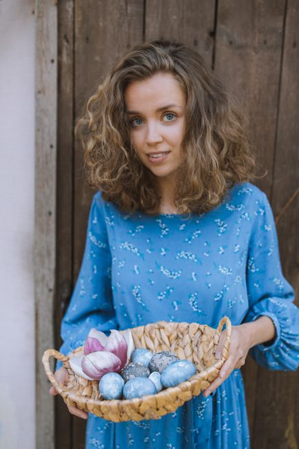 Smiling woman in a blue dress holds a basket of colorful Easter eggs outdoors.