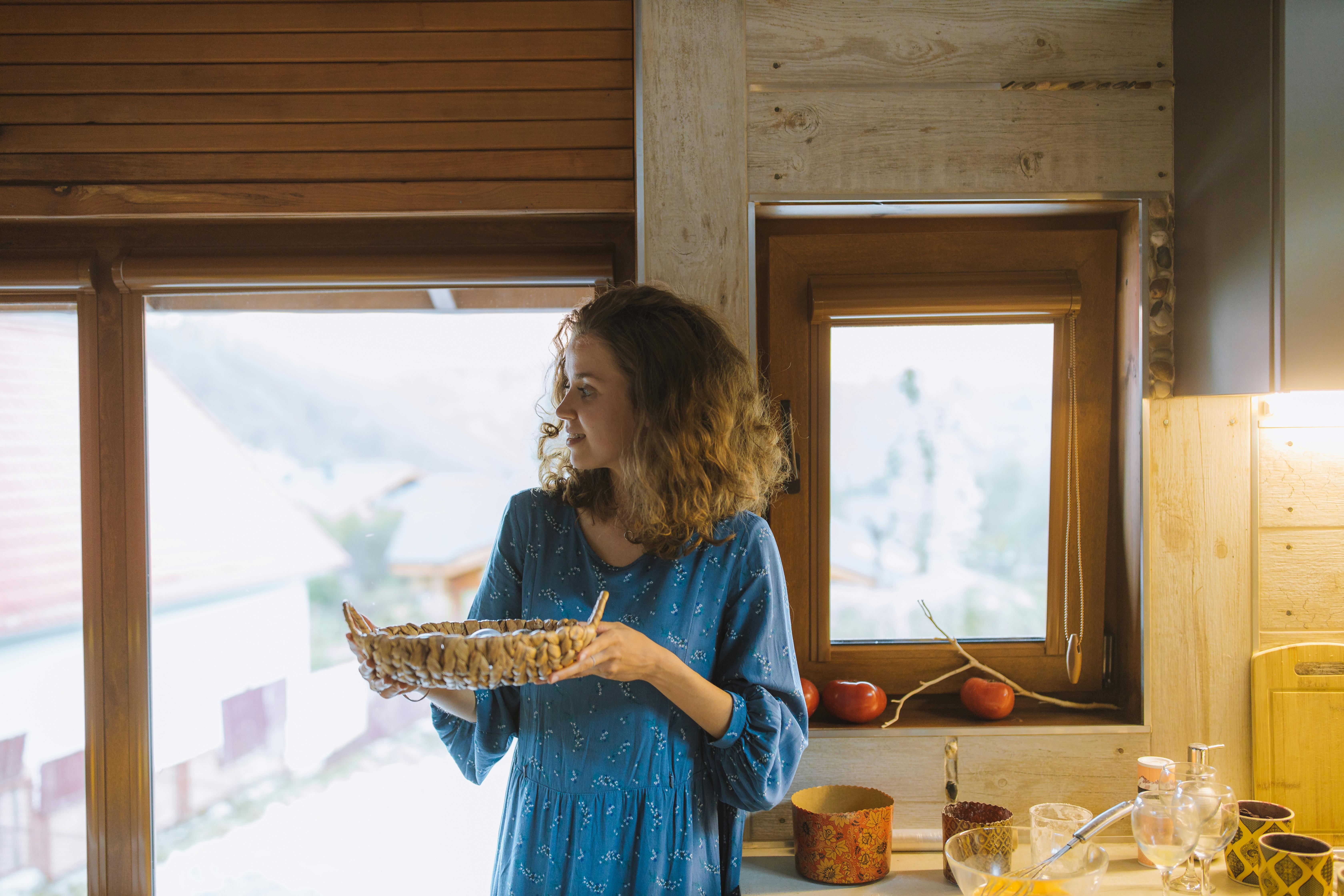 一位身穿蓝色连衣裙的女子，在舒适的厨房里拿着编织篮子，窗外透进自然光