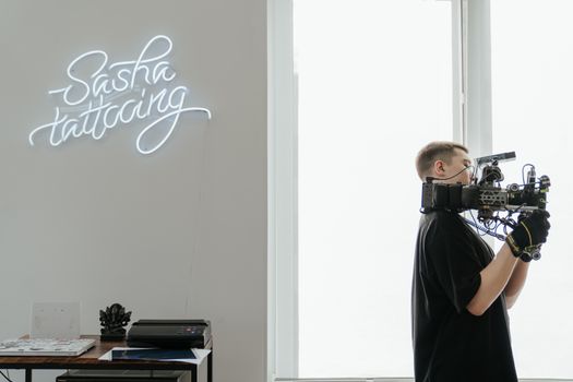 Camera operator filming in a tattoo studio with neon sign 'Sasha Tattooing'.