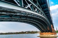 A detailed view of a steel suspension bridge spanning across a calm river under a blue sky.