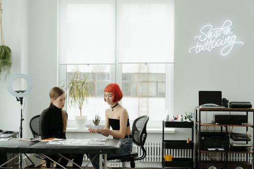 Modern tattoo studio featuring neon lights, an artist with red hair, and a client discussing designs.