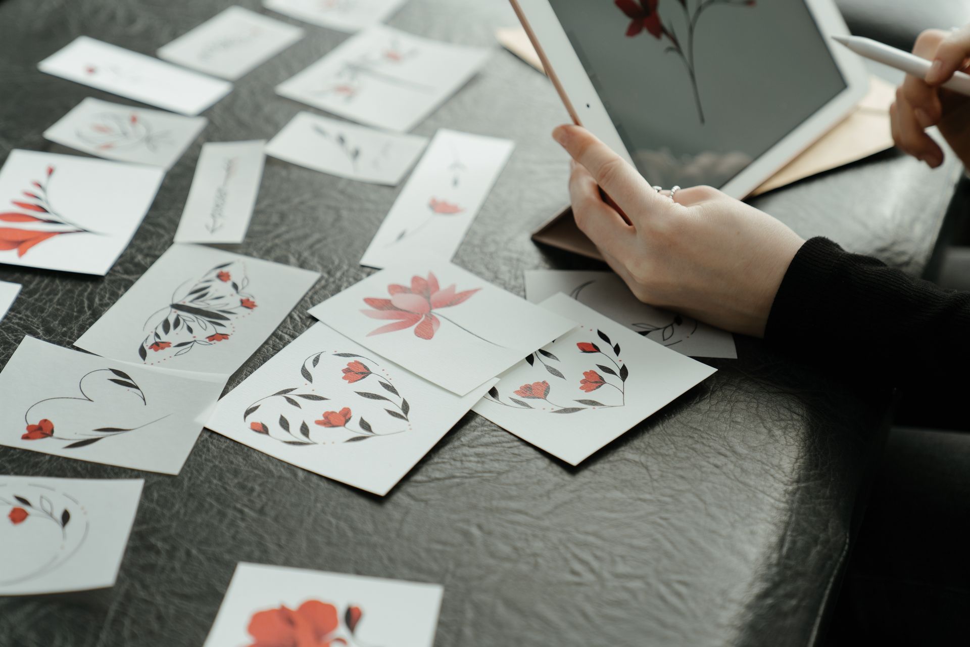 Hands arrange illustrated floral tattoo design cards on a table beside a drawing tablet and stylus.