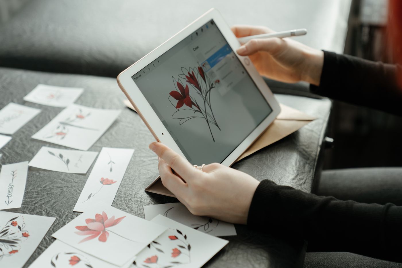 A person uses a stylus on a tablet to refine a floral illustration, with additional botanical drawings spread across a surface.