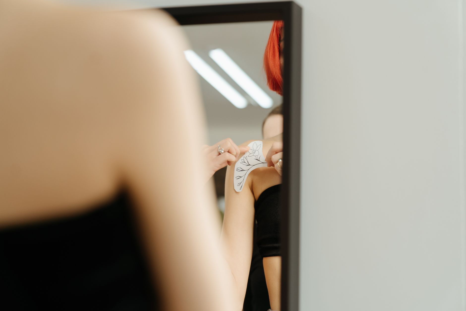 Woman applying a temporary tattoo transfer design to her shoulder in a mirror before committing to permanent ink