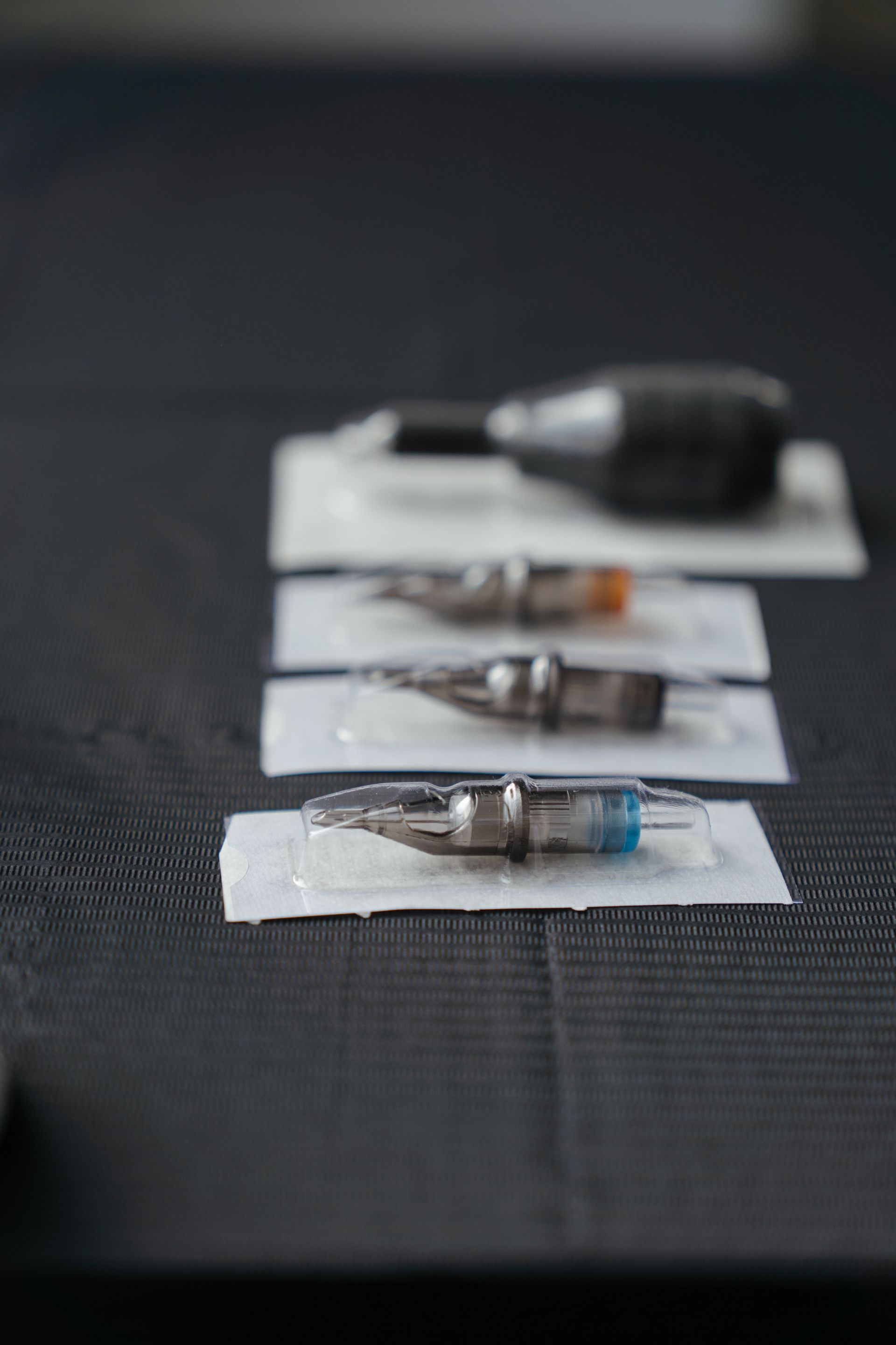 Several individually packaged tattoo needle cartridges are arranged on a dark work surface, with a blurred tattoo machine in the background.