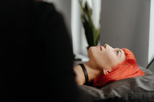 Side profile of a young woman with vibrant red hair lying down in a tattoo studio.