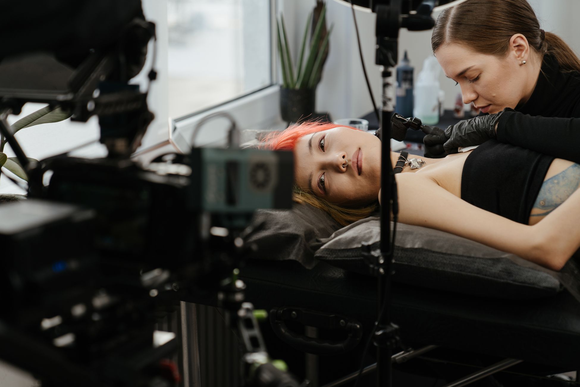 A tattoo artist applies ink in a modern tattoo studio with cameras filming the process.