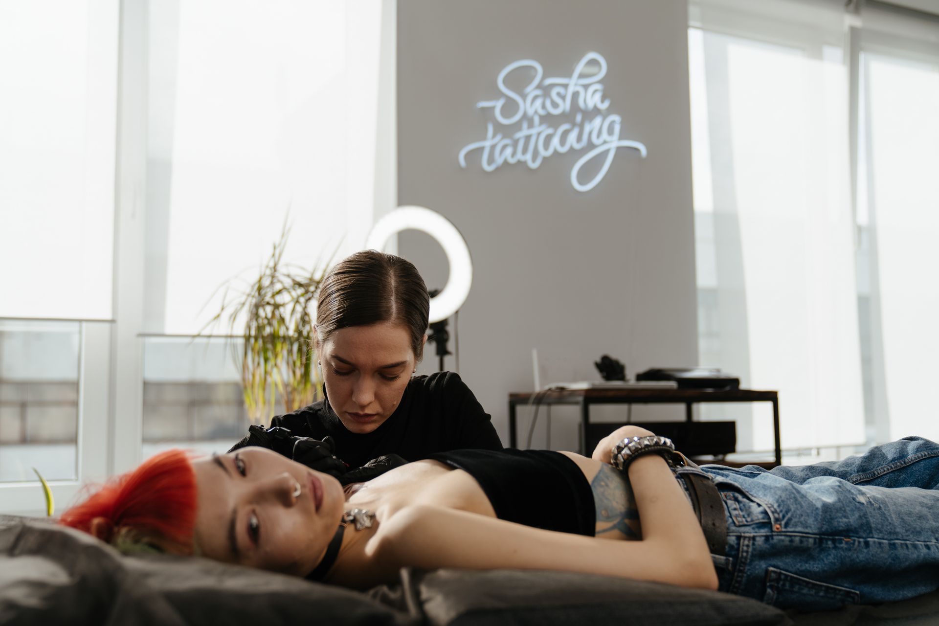 Female tattoo artist creating a design on a client in a stylish studio with neon signage.