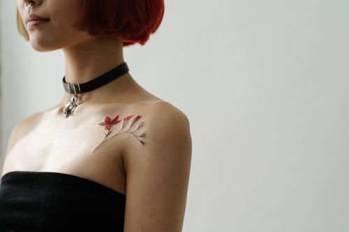 Free An artistic red floral tattoo beautifully adorns a woman's shoulder, captured in a minimalist indoor setting. Stock Photo