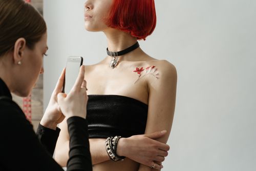 Free A woman with red hair and a shoulder tattoo being photographed by another woman in a tattoo studio. Stock Photo