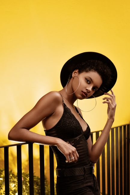 Elegant black woman posing in chic attire with a fedora against a vibrant yellow backdrop.
