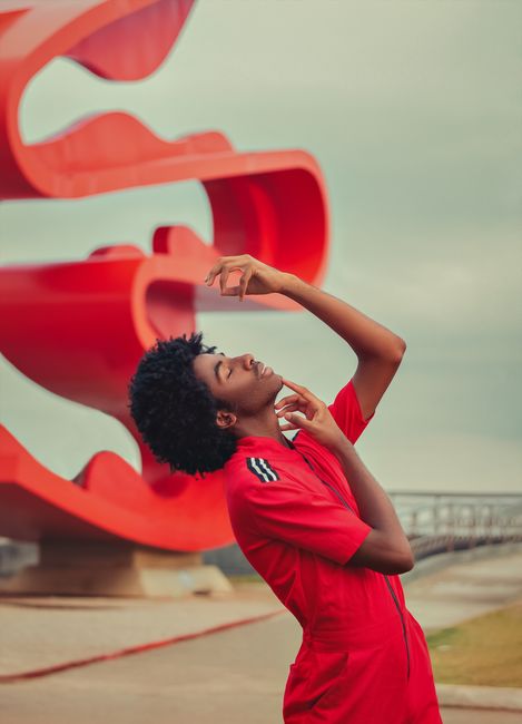 A stylish young man poses expressively outdoors near a striking red sculpture.