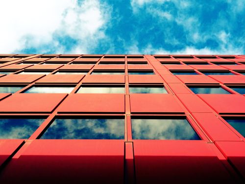 Free Low angle view of a modern red facade building with glass windows reflecting sky and clouds. Stock Photo