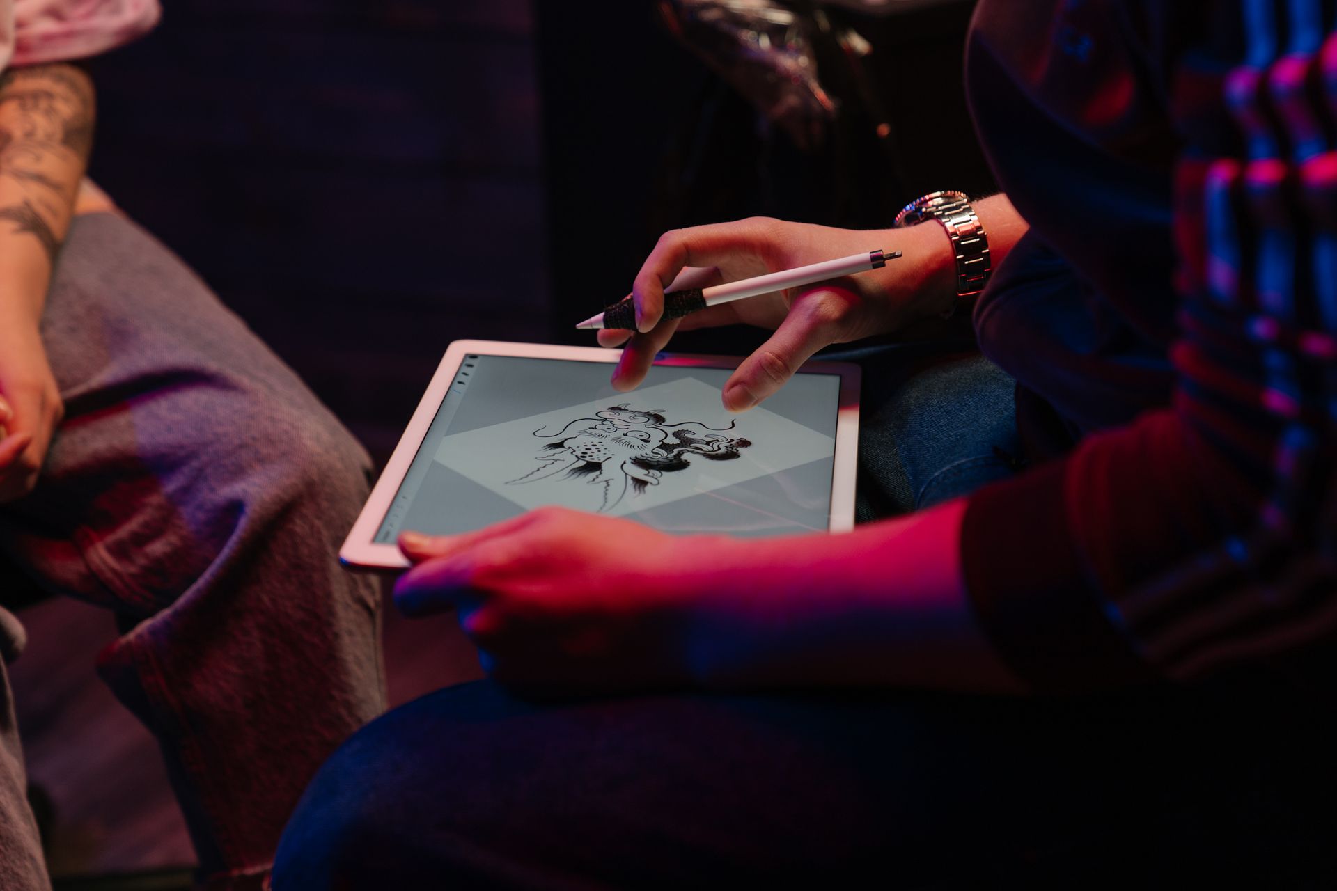 A person uses a stylus on a tablet displaying a tattoo-style illustration while a tattooed person sits nearby under coloured studio lighting.