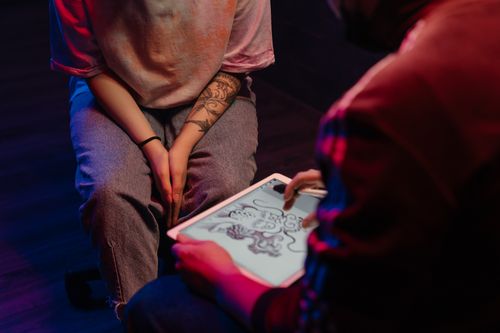 Free A tattoo artist and client discussing a dragon design on a digital tablet in a dimly lit tattoo studio. Stock Photo