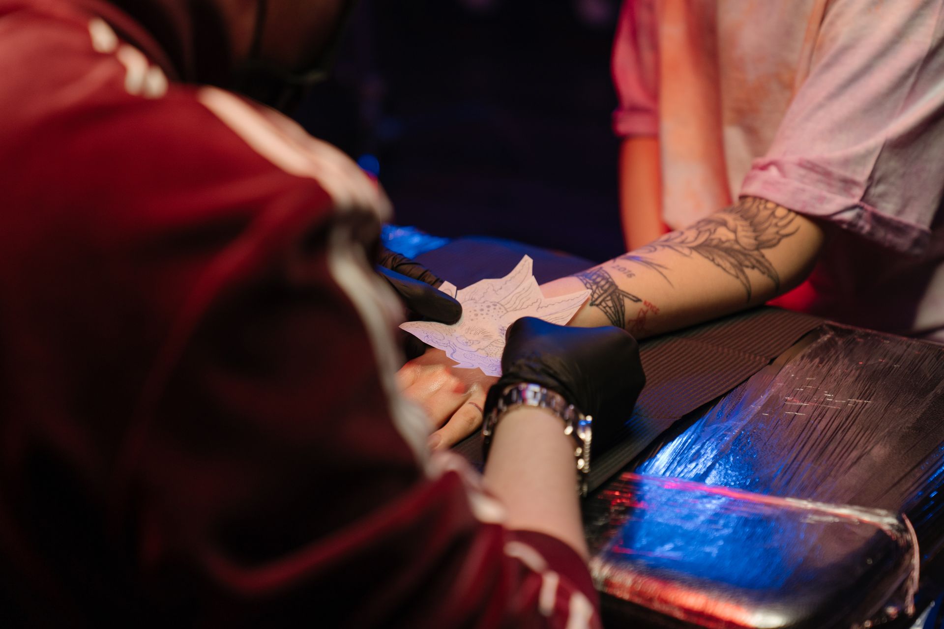 A tattoo artist wearing black gloves applies a paper stencil to a client's tattooed forearm in a dimly lit studio.