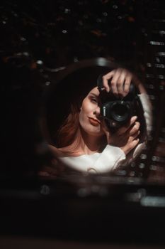 Charming young female with beautiful cheekbones in white shirt taking mirror selfie with photo camera in dark room