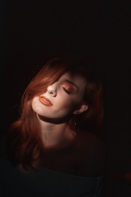 Charming young female with reddish hair and makeup with eyes closed in light of lamp in dark room