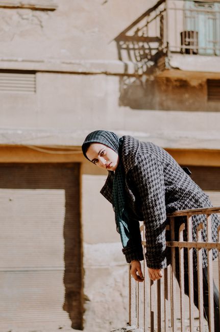 A woman in a coat and headscarf leans on a fence, captured in natural sunlight outdoors.