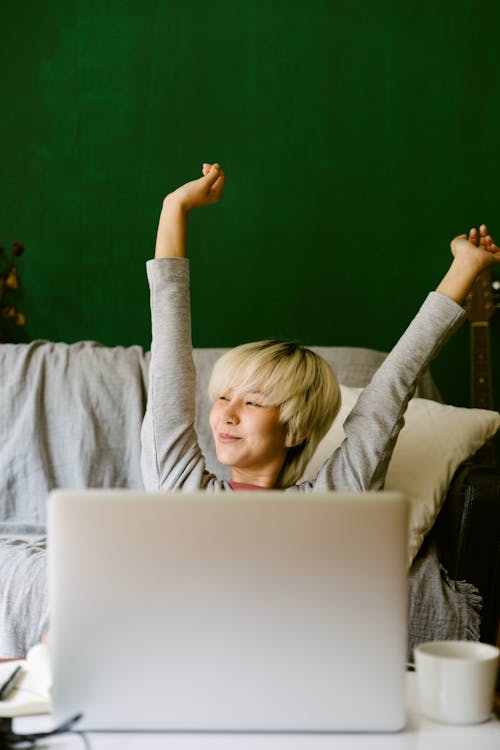 Smiling content young Asian female in casual wear sitting at table at home and using laptop while stretching arm in air and looking away satisfyingly