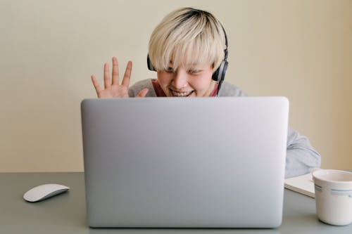 Smiling young Asian female in headphones and casual clothes having conversation via netbook and giving high five while looking at screen and sitting at table with cup of hot drink