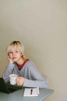 一位穿着休闲装的年轻亚洲女士，正坐在桌前，一边用上网本工作，一边喝着咖啡，目光梦幻地望向米色背景