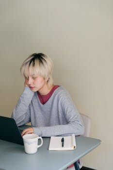 Serious young Asian lady in casual clothes sitting at table with notebook and leaning on hand while working on netbook