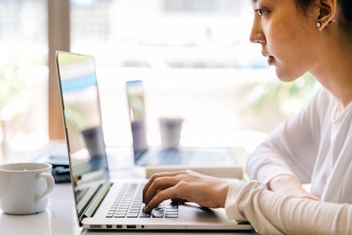 Free Side view of crop serious Asian lady in casual wear sitting at table with cup of beverage and browsing netbook against big glass window on sunny day Stock Photo