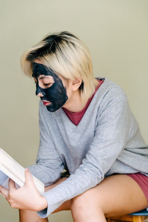 Free Focused female with dyed hair in casual clothes with black facial mask sitting on stool and reading book in daylight on beige background Stock Photo
