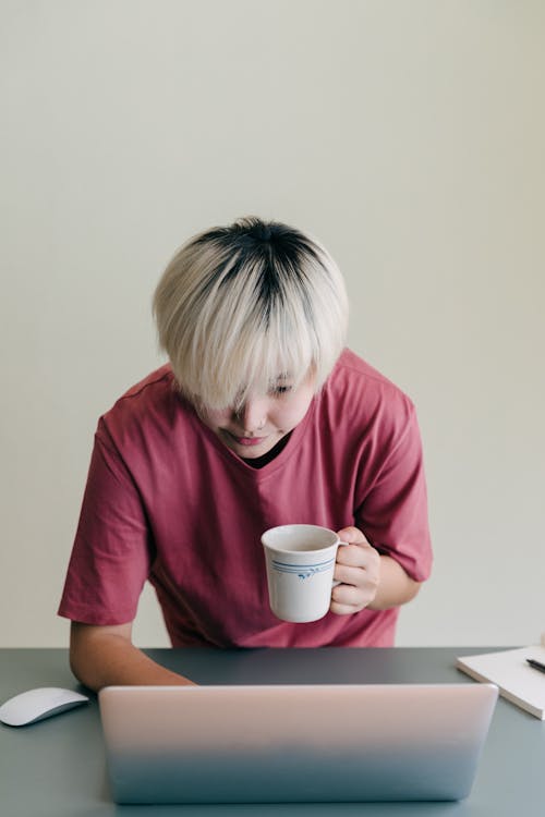 Serious young Asian lady in casual shirt with cup of hot drink typing on netbook while bending above table and working remotely on project on beige background in daylight