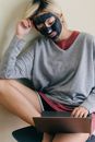 Young woman with cosmetic facial mask sitting on chair while using laptop at home