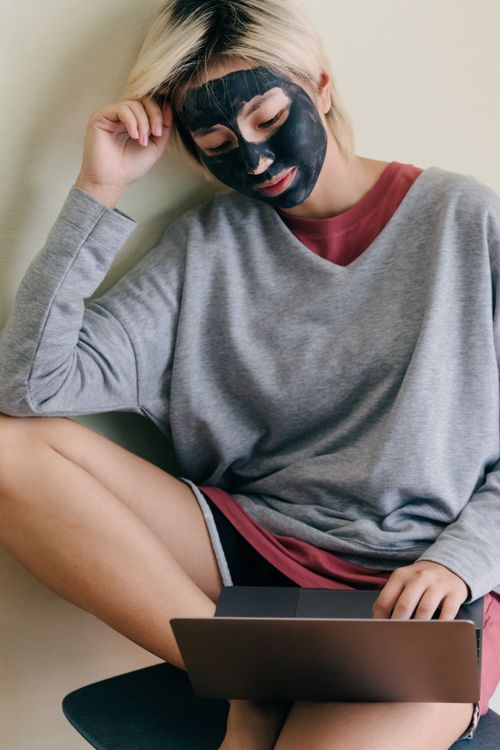 Free Young Asian female in casual wear with black cosmetic facial mask sitting relaxed on chair leaning on hand while using laptop placed on knee at home Stock Photo