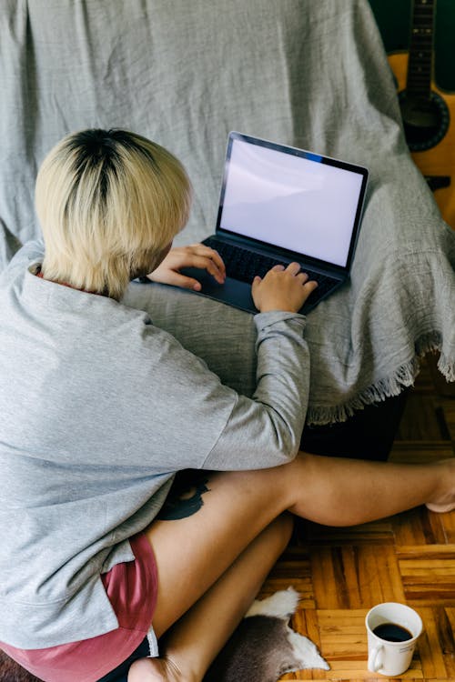 Free Back view of faceless young female in casual wear sitting on floor with cup of coffee and using laptop placed on armchair Stock Photo