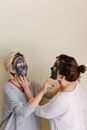Playful girlfriends with facial mud mask touching chins of each other on beige background
