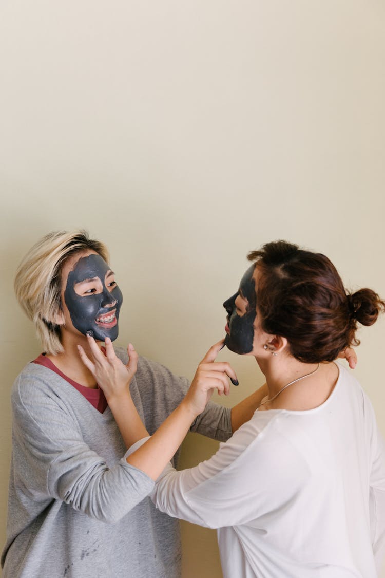 Playful Girlfriends With Facial Mud Mask Touching Chins Of Each Other On Beige Background
