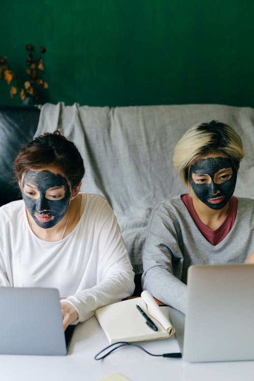 From above of serious young Asian girlfriends in casual clothes with black facial mud masks sitting at table while using laptops at home
