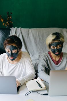 一位严肃的年轻亚洲女友穿着休闲装，戴着黑色口罩，在家里的桌旁用着笔记本电脑