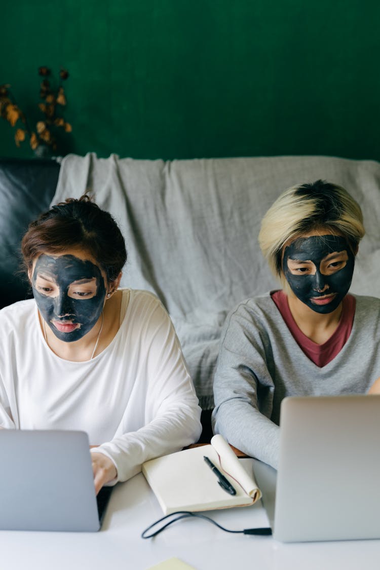 Focused Young Women With Black Facial Masks Using Laptops At Home