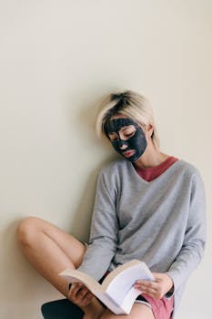 Concentrated young Asian female in casual wear with black cosmetic facial mask sitting relaxed on chair while reading book at home