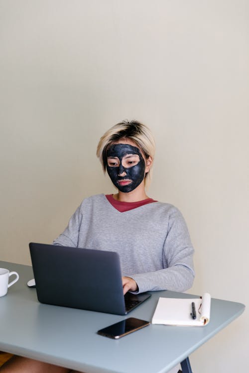 Free From above of concentrated young ethnic female student in casual outfit with black clay face mask using laptop while sitting at table on beige background Stock Photo