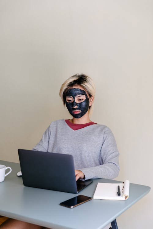 From above of concentrated young ethnic female student in casual outfit with black clay face mask using laptop while sitting at table on beige background