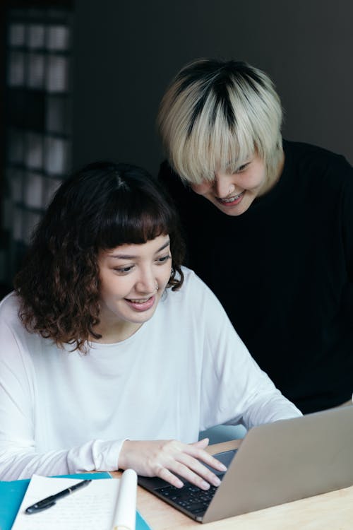 Positive ethnic women smiling while using laptop and browsing information while working on project together at workplace during meeting looking at screen