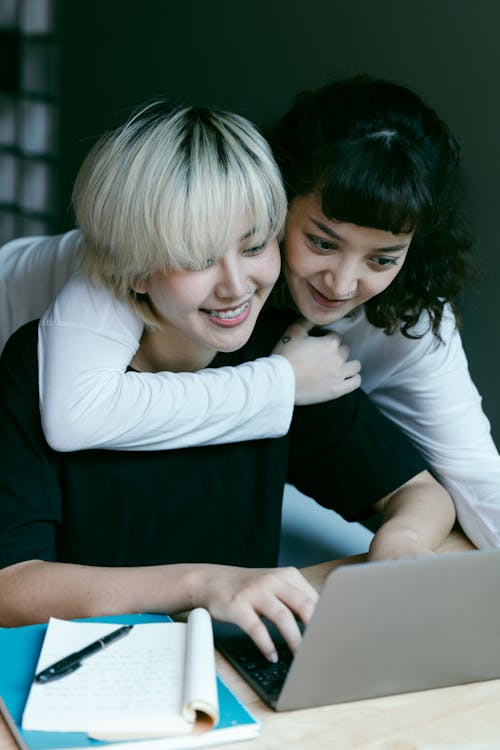 Positive ethnic woman with laptop smiling while sitting at table and working on project while coworker friend hugging and looking at screen