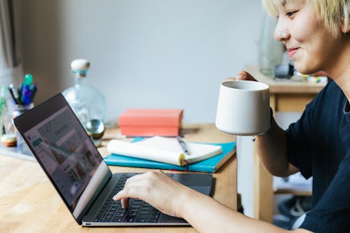Free Side view of cheerful Asian female using laptop while sitting at table with cup of hot drink Stock Photo